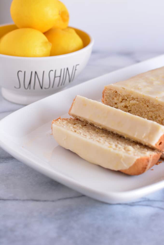 Glazed lemon loaf sliced on a white place with a bowl of lemons in the background